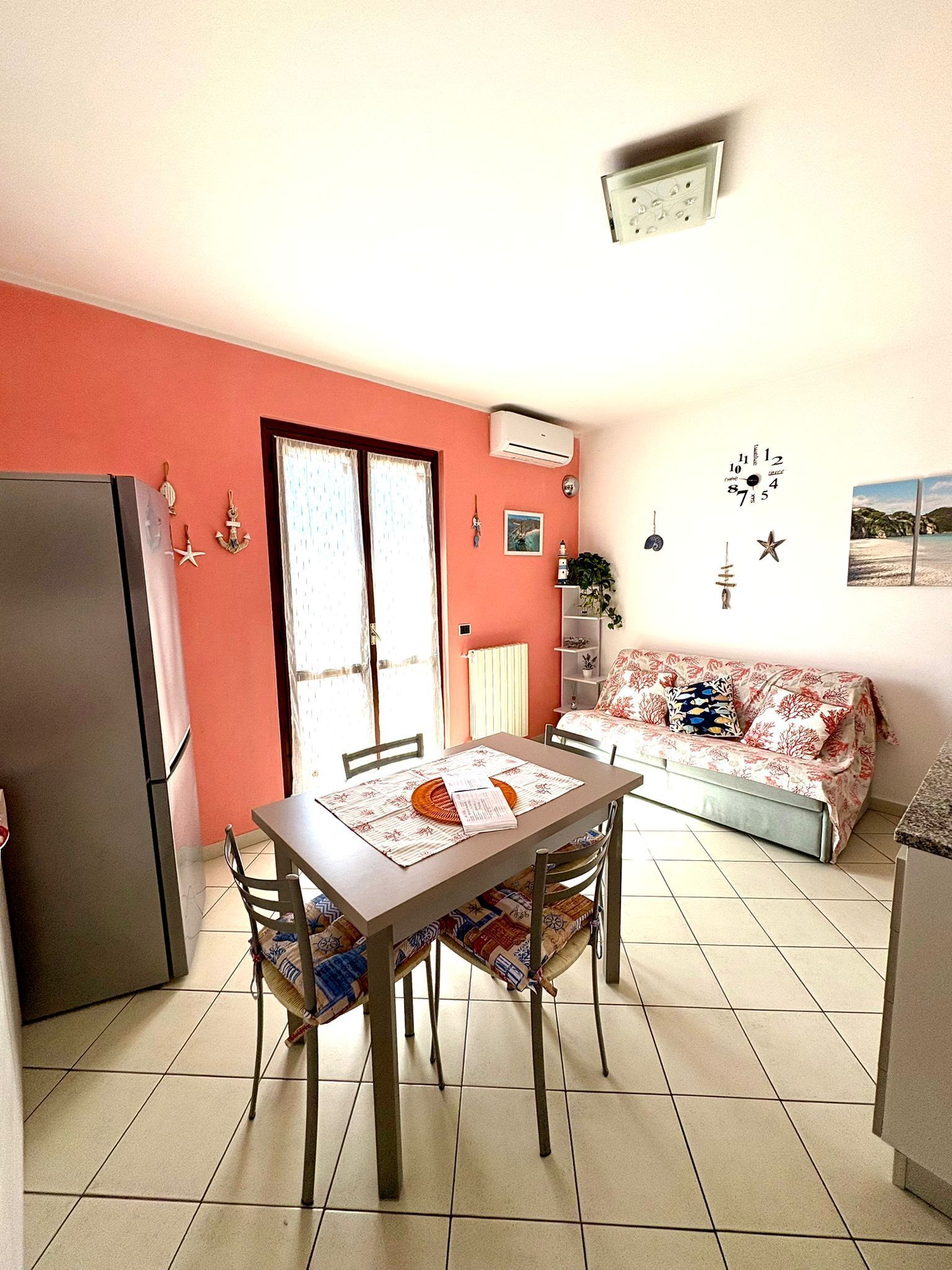A compact living space with a dining table, chairs, a sofa, a refrigerator, and a window with curtains.