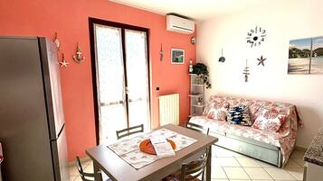 A compact living space with a dining table, chairs, a sofa, a refrigerator, and a window with curtains.