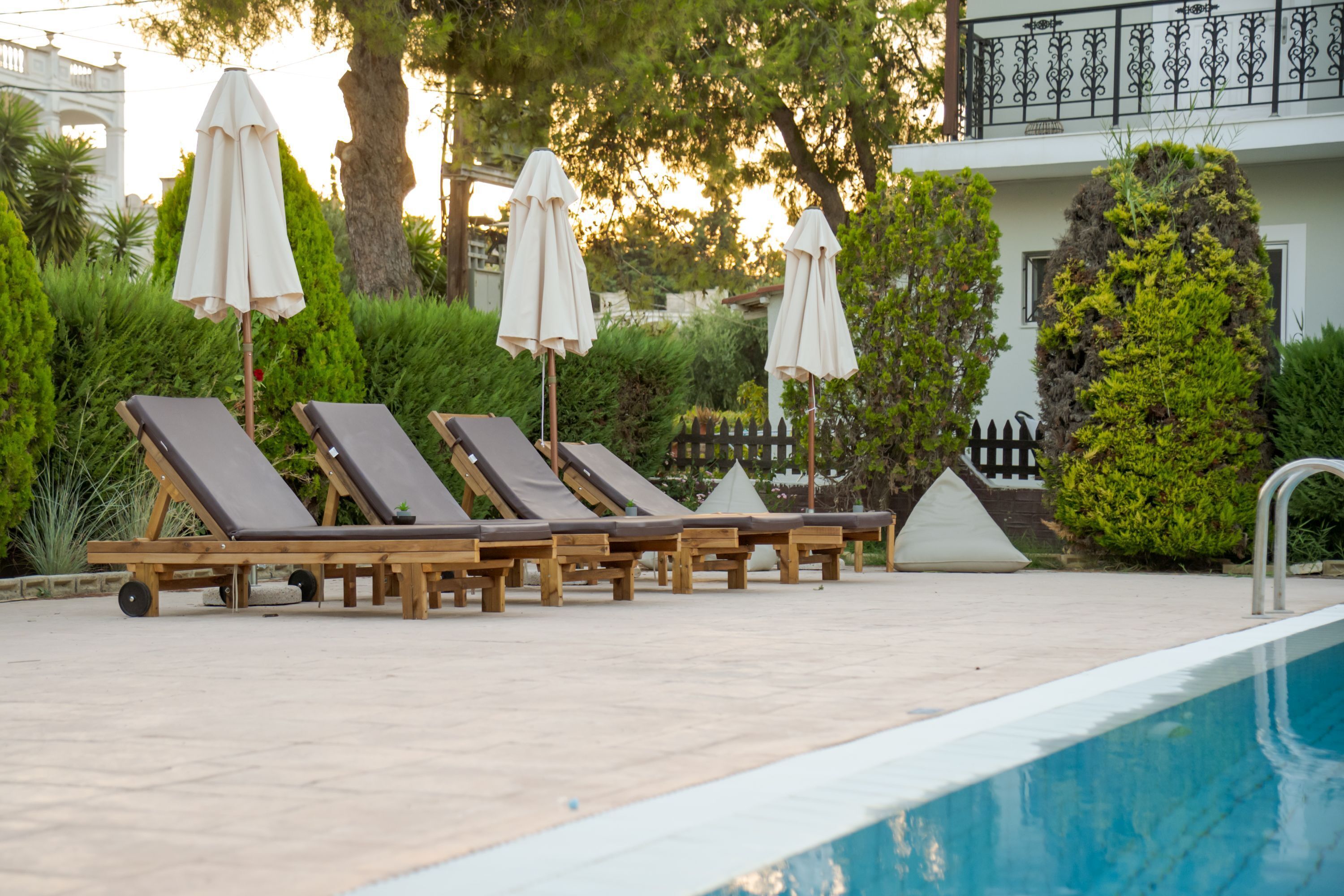 Un espace aménagé au bord de la piscine, avec des chaises longues et des parasols, entouré de verdure et bordé d’un bâtiment doté d’un balcon.