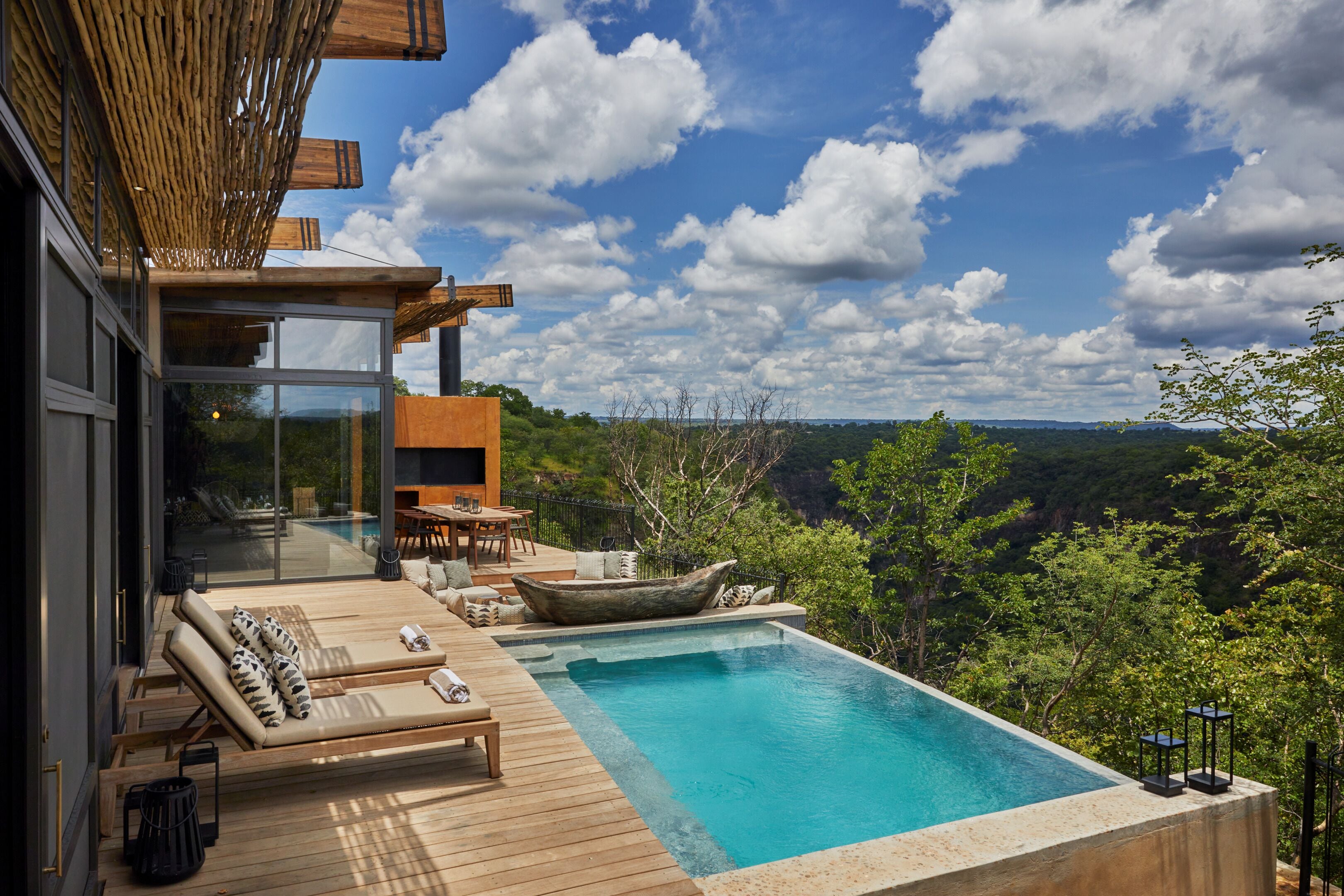 A modern outdoor pool area with wooden deck, lounge chairs, and a view of the surrounding forest.