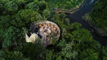Une vue aérienne d’une structure circulaire en bois, dotée d’un espace extérieur aménagé pour s’asseoir, et entourée d’une forêt dense et d’une rivière.