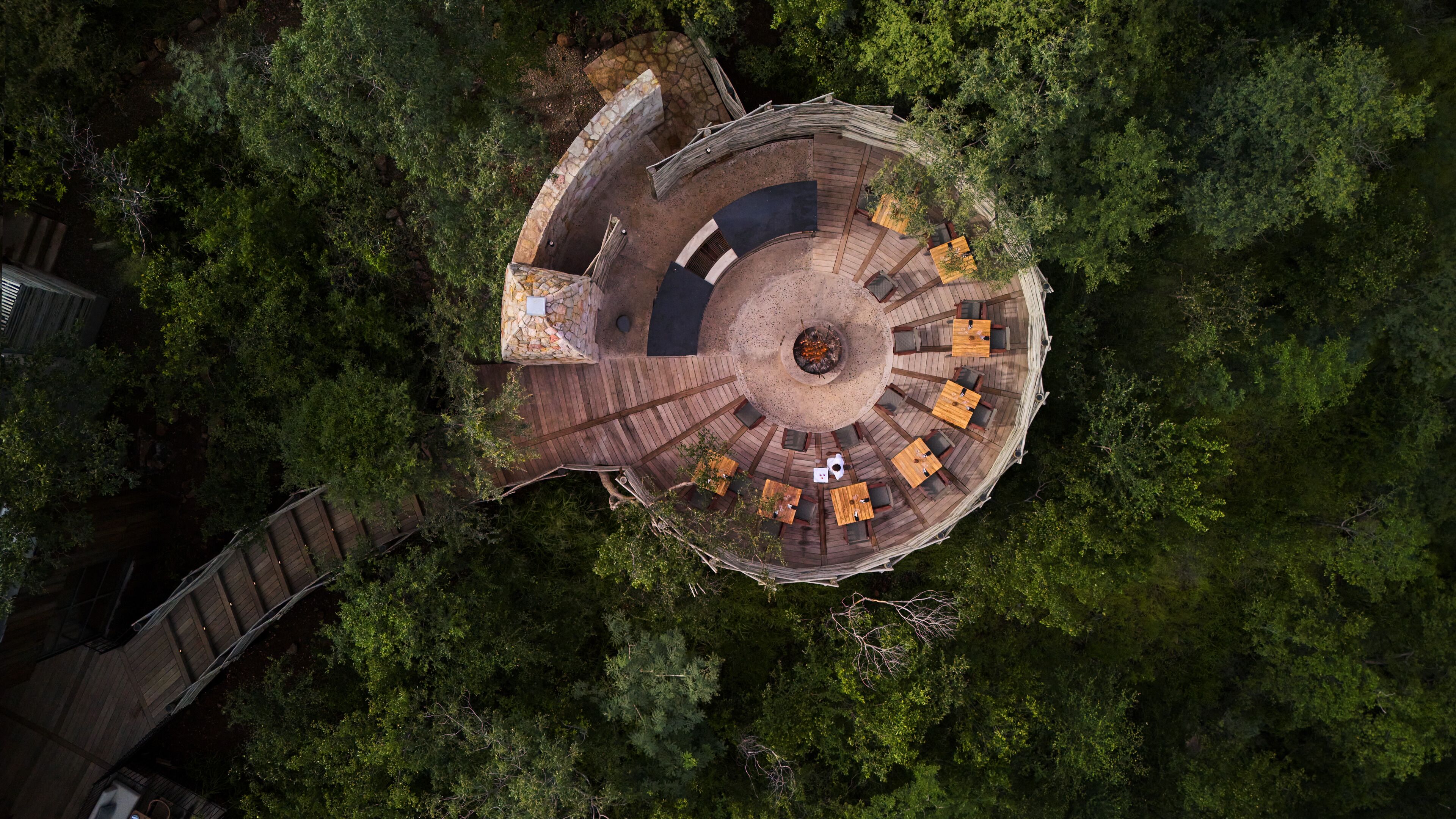 An aerial view of a circular wooden deck with seating around a central fire pit, surrounded by dense forest.