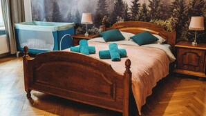 A wooden bed with a headboard, two nightstands, a blue crib, and a forest-themed wall mural. - FatCat Apartments (Kraków)