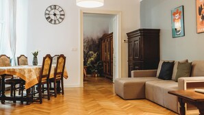 A dining area with a wooden table and chairs, a clock on the wall, and a sofa in the living room. - FatCat Apartments (Kraków)
