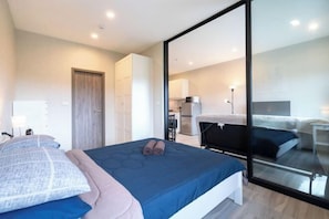 A modern bedroom with a large bed, a glass door leading to a balcony, and a visible kitchen area.
