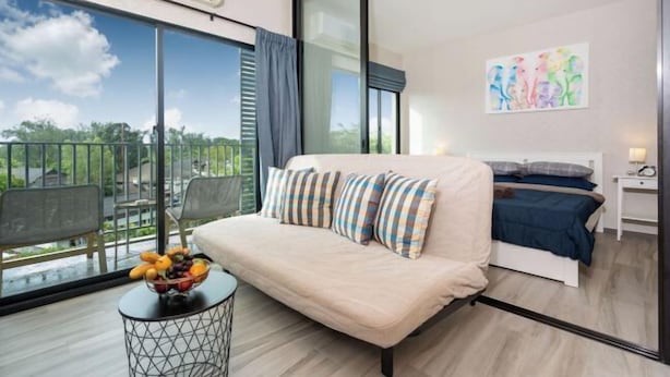 Een moderne slaapkamer met een slaapbank, een klein rond tafeltje met fruit, een bed met een blauwe dekbedovertrek en een groot raam met uitzicht op groen.