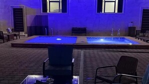 Uma área de piscina ao ar livre com um edifício iluminado em azul ao fundo, uma área de estar com cadeiras e uma mesa com tampo de vidro.