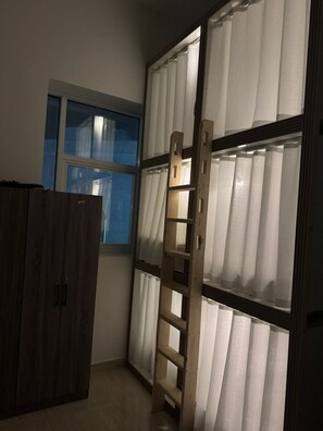 A room with a wooden wardrobe, a window with blue trim, and a wooden ladder leaning against a wall with white curtains. - Travel Hub Near Business Bay By Al Taj (Dubai)