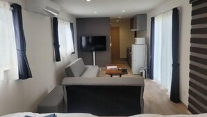 A modern hotel room with a sofa, a flat-screen TV, a small kitchen area, and a large window with curtains. - Toya Center Village (Toyako)
