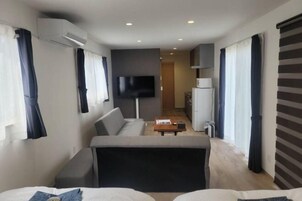Una habitación de hotel moderna con sofá, televisor de pantalla plana, pequeña cocina y un amplio ventanal con cortinas.
