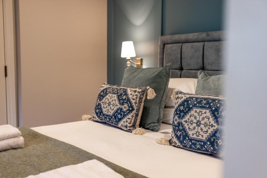 Un lit bien fait, agrémenté d’oreillers à motifs, d’une lampe de chevet et d’un plaid vert.