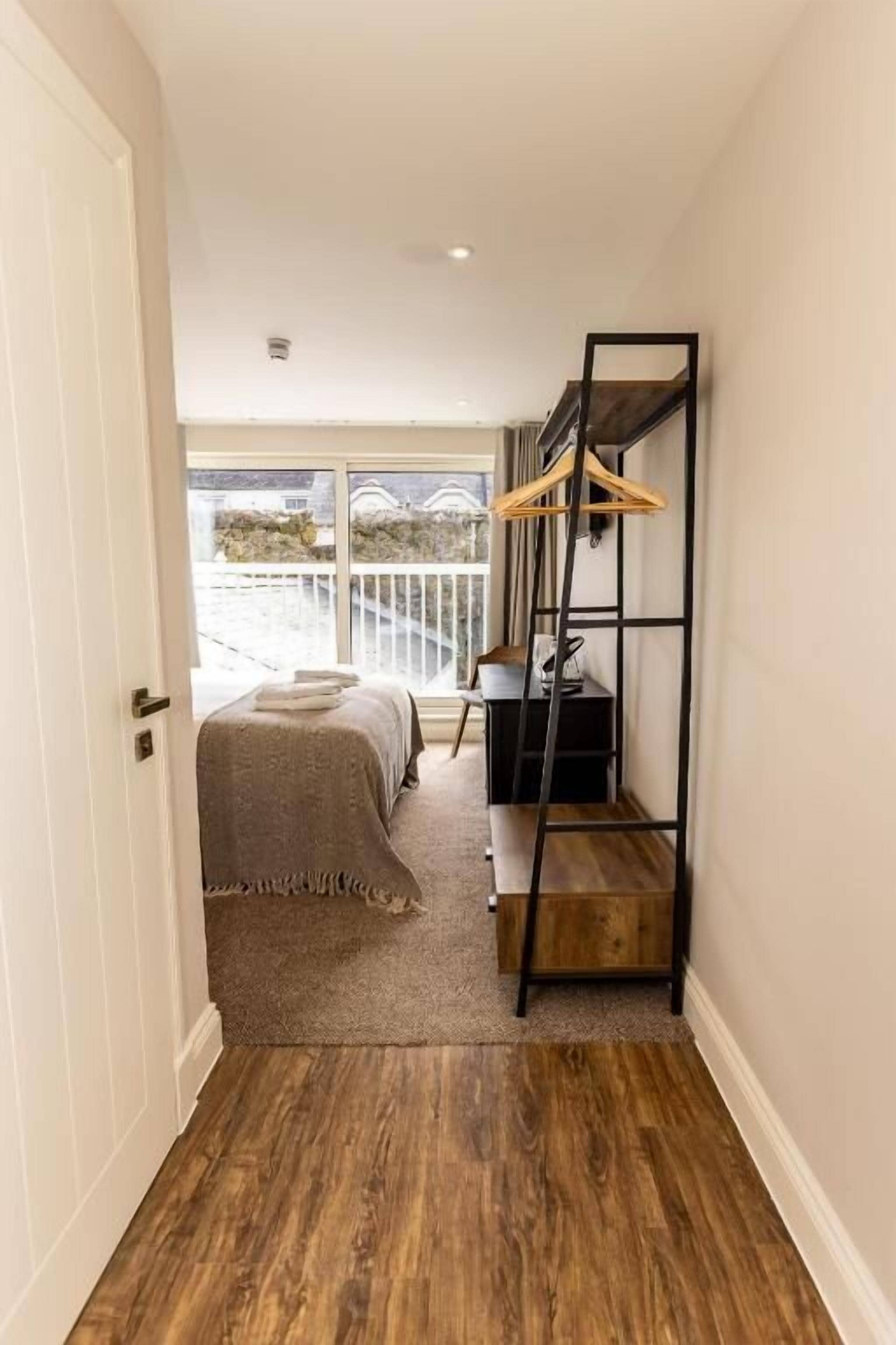 Un couloir étroit, doté d’un plancher en bois, mène à une chambre à coucher qui comprend un lit et un balcon. Une étagère en métal noir, sur laquelle est fixé un cintre en bois, est fixée au mur.