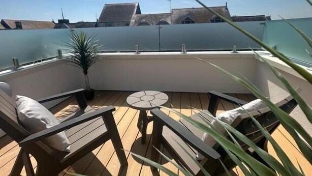 A wooden deck with a round table and two lounge chairs, overlooking a body of water and buildings. - The Town Crier (Tenby)