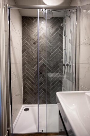 Una ducha a ras de suelo con patrón de mosaico de espiga, puerta de cristal y lavabo blanco.