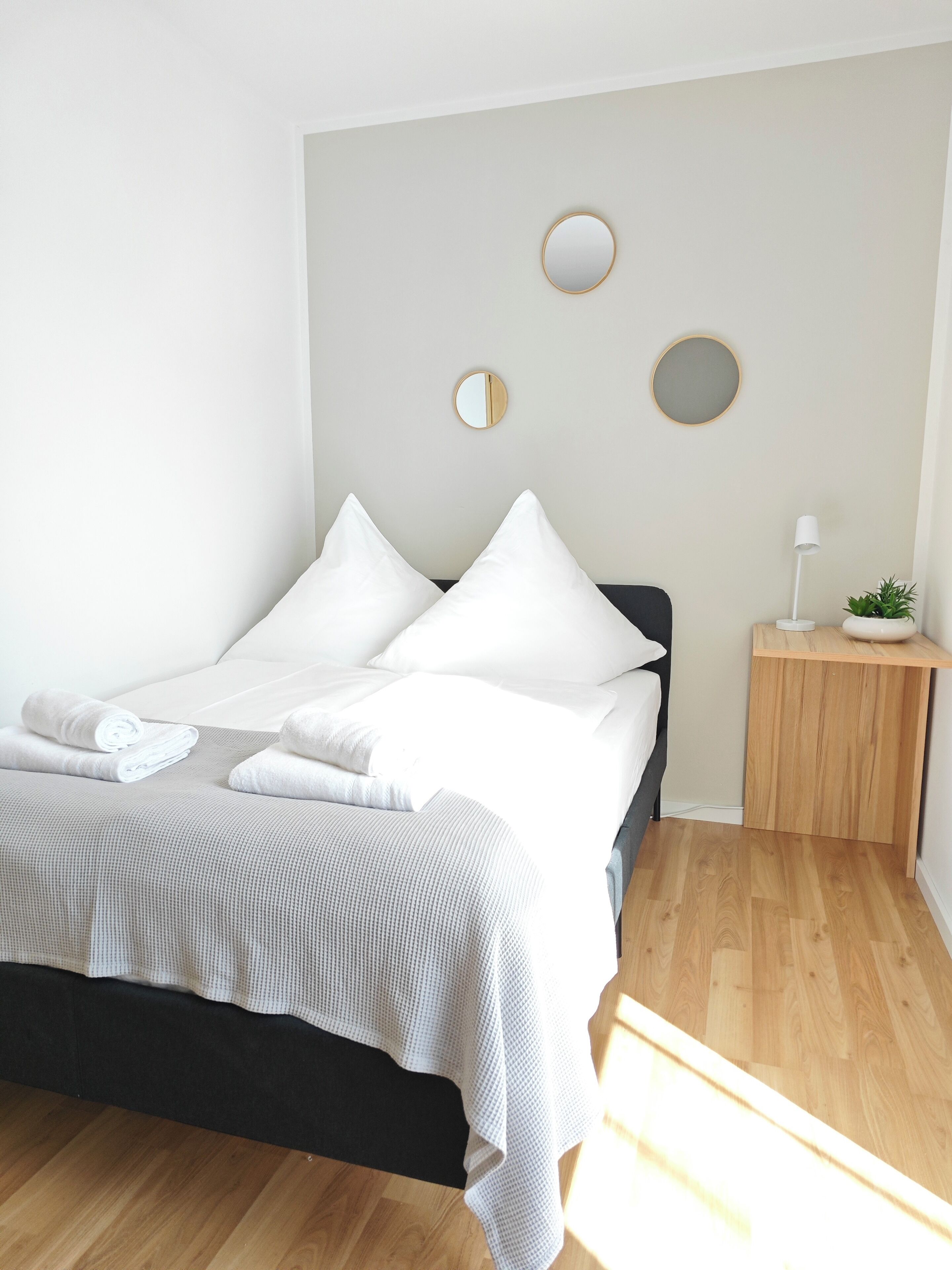 Una cama bien hecha con sábanas y almohadas blancas, una mesita de noche con una lámpara y una planta pequeña, y dos espejos colgados en la pared.