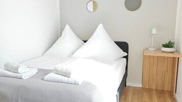Una cama bien hecha con sábanas y almohadas blancas, una mesita de noche con una lámpara y una planta pequeña, y dos espejos colgados en la pared.