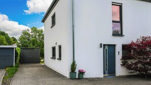 Ein modernes Gebäude mit flachem Dach, einer Tür und Fenstern. Übertöpfe mit Pflanzen und ein gepflasterter Zugangsweg sind ebenfalls vorhanden.