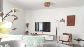 Ein modernes Wohnzimmer mit Flachbildfernseher, Korbmöbeln und einem Essbereich mit Glastisch und Tellern.