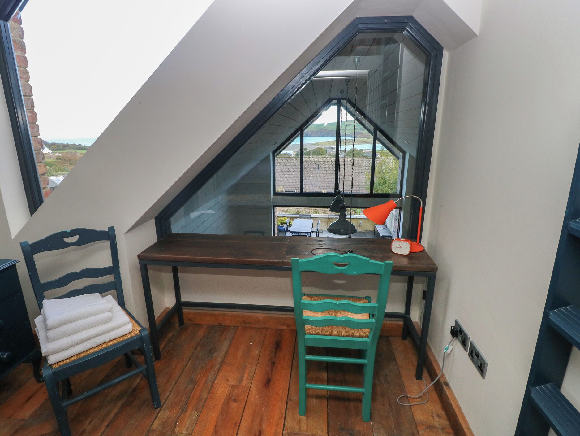 Un dormitorio estilo altillo pequeño con techo inclinado, un escritorio de madera, una silla, una lámpara y vistas al exterior.