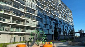 Un immeuble moderne à plusieurs étages, doté de nombreux balcons, d’une aire de jeux avec une structure d’escalade verte et de coins salon orange.