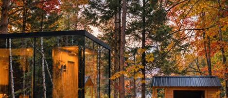 A glass-enclosed cabin with a hot tub, wooden deck, and outdoor seating area surrounded by autumn foliage.
