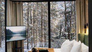Quarto moderno com cama grande, televisão de tela plana, mesa de centro e vista para uma floresta nevada através de amplas janelas.