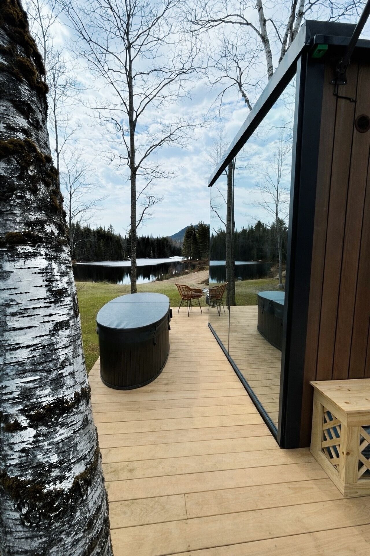 Une terrasse en bois dotée d’un jacuzzi, de chaises et d’une véranda fermée par des baies vitrées, offrant une vue sur un lac et une forêt.