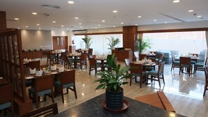A spacious hotel lobby with multiple dining areas, wooden furniture, and potted plants. - DEVENTURE HOTEL (New Delhi)