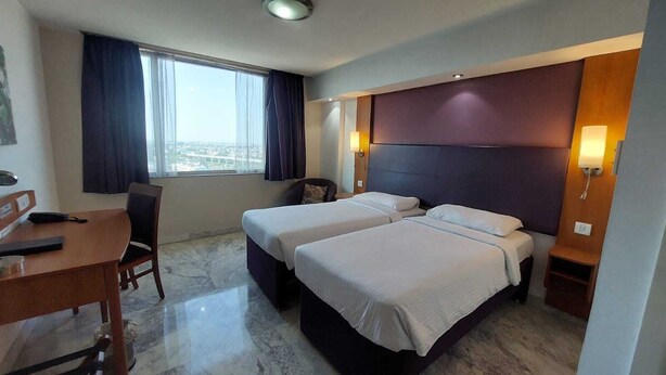 A hotel room with two beds, a desk, and a chair. There is a window with curtains, a view of the city, and a wall-mounted lamp. - DEVENTURE HOTEL (New Delhi)