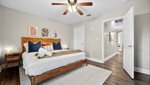 A bedroom with a wooden bed, white bedding, a ceiling fan, and a hallway leading to another room. - Walk to AT&T Stadium | Modern 2BR With Backyard (Arlington)