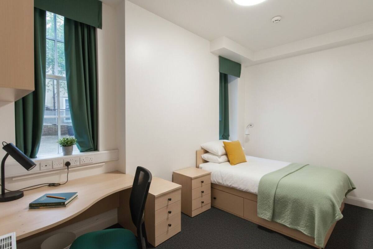 A bedroom with a desk, a bed with a green blanket, a chair, and a window with curtains.