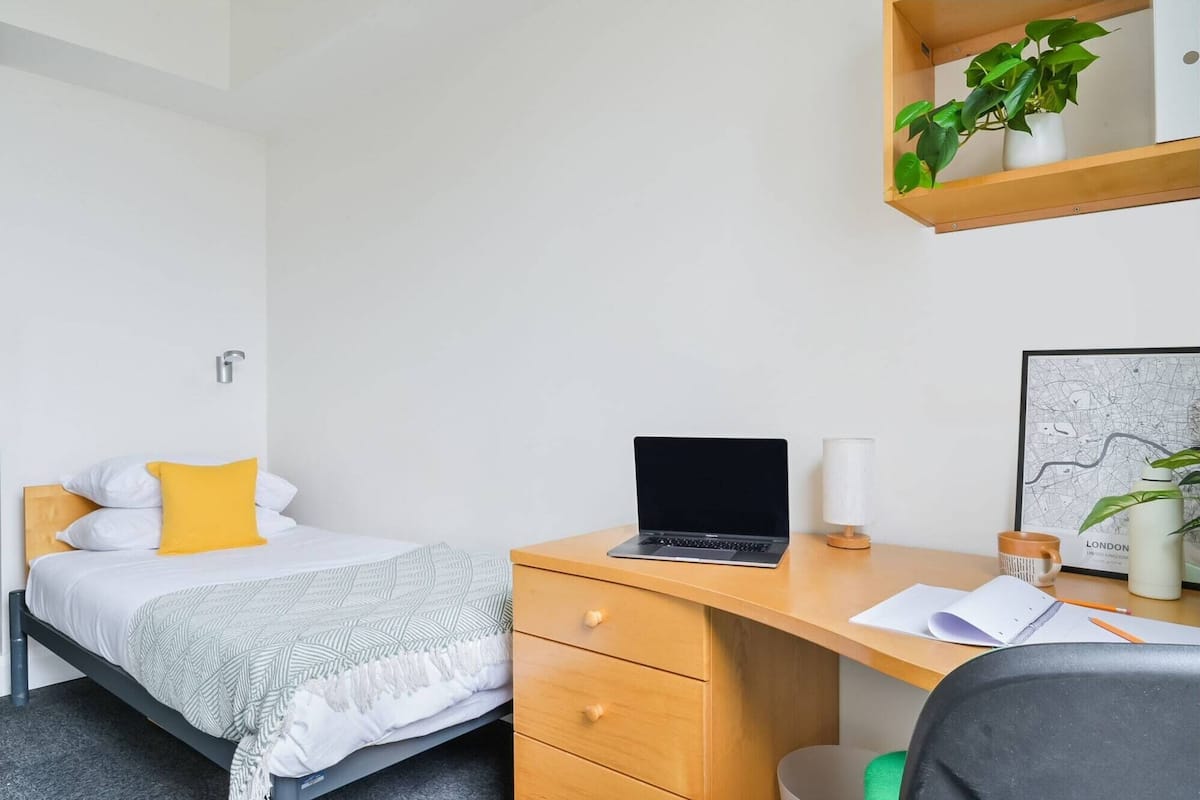 A bedroom with a bed, a desk, a laptop, a map, and a plant.