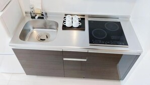 A compact kitchen with a stainless steel sink, a black induction cooktop, and a small cabinet with a tray of white cups. - WANO (Osaka)