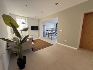 A modern living room with a sofa, a coffee table, a large potted plant, a flat-screen TV mounted on the wall, and a dining area with chairs and a table.