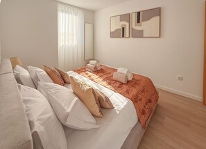 Un dormitorio con cama, almohadas, una colcha, una ventana con persianas y dos cuadros en la pared.