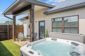 A hot tub in a backyard with a covered patio, a wooden fence, and a house with large windows.
