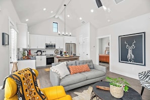 A modern living room with a grey sofa, yellow armchair, and a dining area with white cabinets and a kitchen in the background.