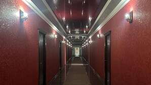 Un pasillo largo y estrecho con paredes rojas y suelo oscuro, flanqueado por puertas e iluminado por luces del techo.