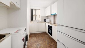 A compact kitchen with white cabinets, a washing machine, a microwave, and a tiled floor. - Beachfront Bliss 2BR Ocean Breeze Hideaway,Parking (Surfers Paradise)