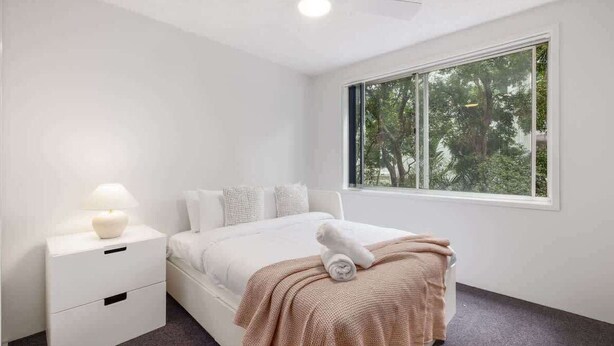 A bedroom with a bed, a nightstand, a lamp, and a window with a view of trees. - Beachfront Bliss 2BR Ocean Breeze Hideaway,Parking (Surfers Paradise)