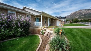 A two-story house with a front porch, a stone facade, and a well-maintained lawn. - Grand Ogden Lodge, Hot Tub, Close to Ski Resorts – 6 BR | 5 Baths | Hot Tub (North Ogden)
