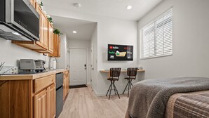 A compact room with a kitchenette, a bed, a TV displaying streaming services, and a window with blinds. - Grand Ogden Lodge, Hot Tub, Close to Ski Resorts – 6 BR | 5 Baths | Hot Tub (North Ogden)