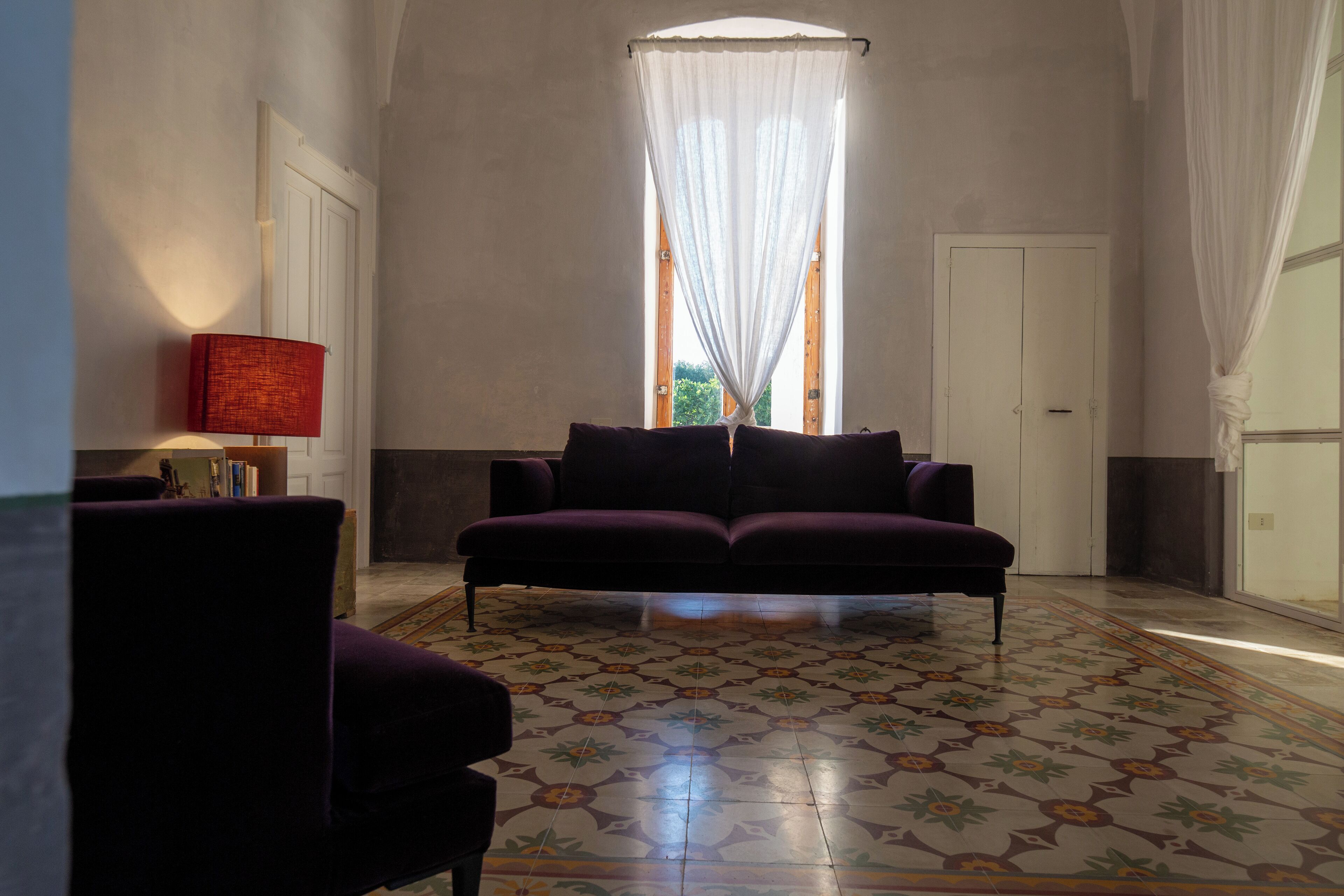 A room with a patterned floor, a purple sofa, a red lamp, and a window with sheer curtains.