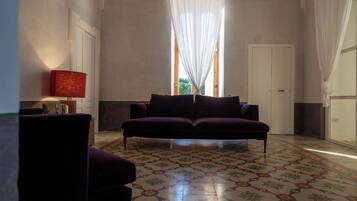 Una habitación con piso estampado, un sofá morado, una lámpara roja y una ventana con cortinas translúcidas.