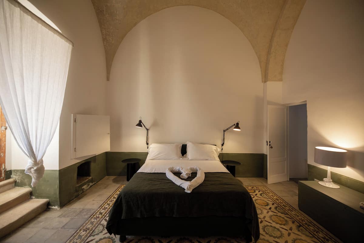 A bedroom with a bed, two bedside lamps, a nightstand, a dresser, a patterned rug, and a window with curtains.