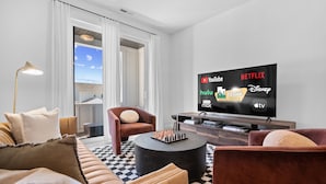 Un salon moderne doté d’un grand téléviseur à écran plat diffusant divers services de streaming, d’un canapé confortable, d’une table basse et offrant une vue sur l’extérieur grâce à de grandes fenêtres.