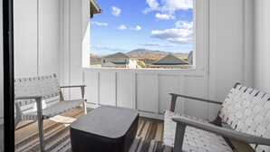 Un balcon avec deux fauteuils en osier blanc, un pouf noir et une vue sur des maisons et des montagnes au loin.