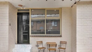 Entrada de un edificio con una puerta negra, una ventana que da a un edificio de ladrillo, y tres sillas de madera con una mesa afuera.