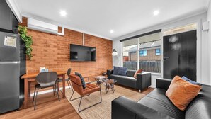 Una sala de estar moderna con pared de ladrillo, un televisor de pantalla plana, una zona de comedor con mesa y sillas, y un sofá con cojines.