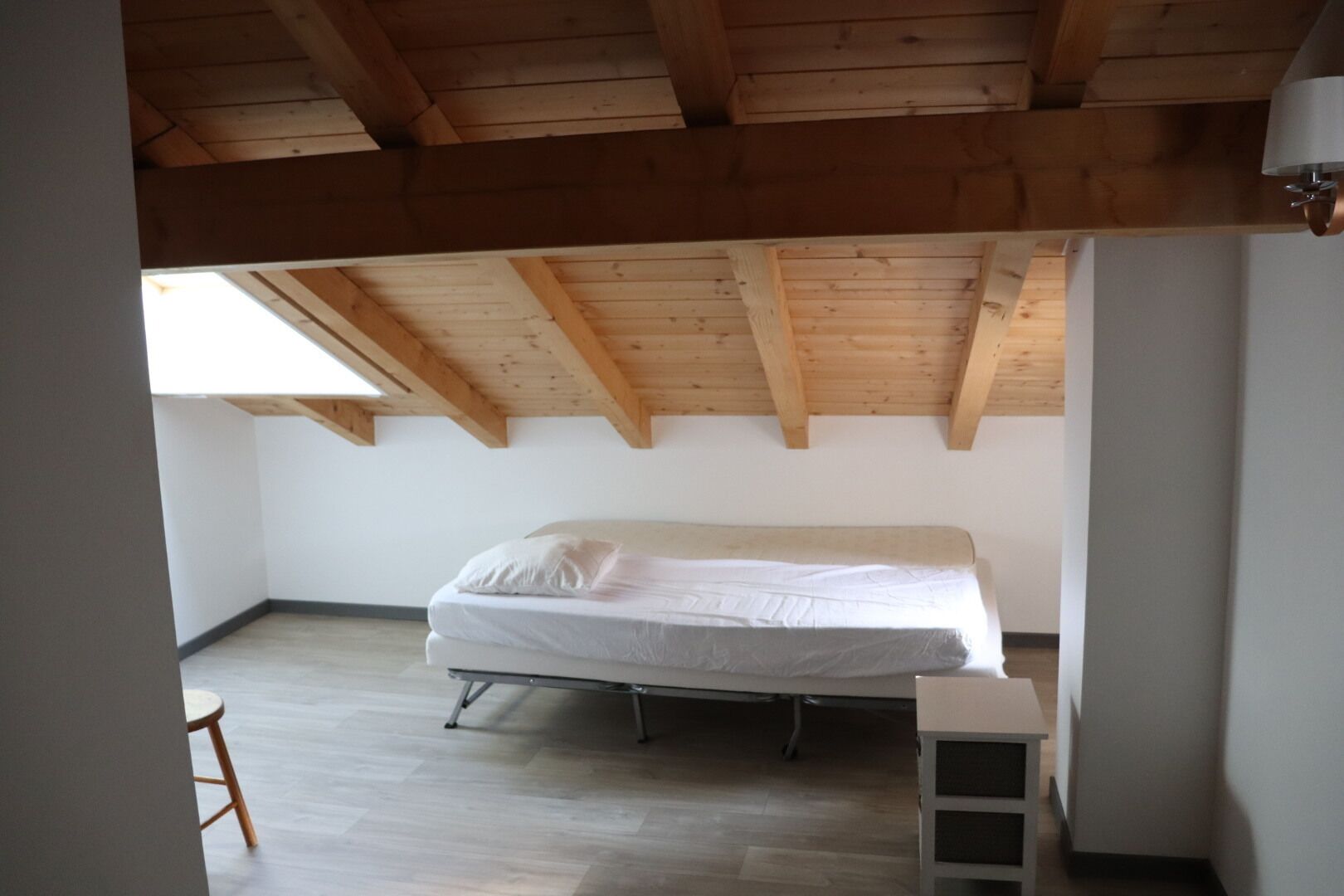 Pequeña habitación con cama individual, techo inclinado y vigas de madera, una mesita y una escalera.